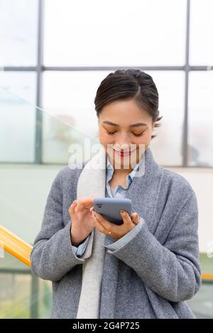 Junge schöne asiatische Geschäftsfrau hält ein Mobiltelefon in einem Bürogebäude, um Nachrichten zu senden und Social Media zu durchsuchen Stockfoto