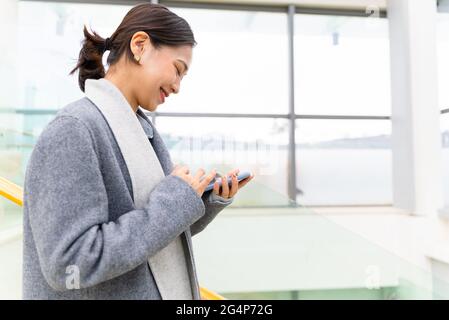 Junge schöne asiatische Geschäftsfrau hält ein Mobiltelefon in einem Bürogebäude, um Nachrichten zu senden und Social Media zu durchsuchen Stockfoto