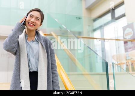 Low-Angle-Aufnahme einer jungen, schönen asiatischen Geschäftsfrau, die mit einem Mobiltelefon im Bürogebäude mit Kopierfläche telefoniert Stockfoto