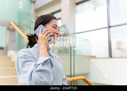 Low-Angle-Aufnahme einer jungen, schönen asiatischen Geschäftsfrau, die mit einem Mobiltelefon im Bürogebäude mit Kopierfläche telefoniert Stockfoto