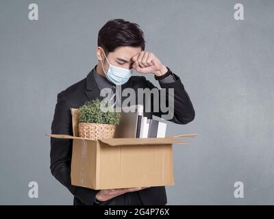 Der junge Geschäftsmann wird gefeuert, hält einen Karton mit Büromaterial und steht vor einer grauen Wand Stockfoto