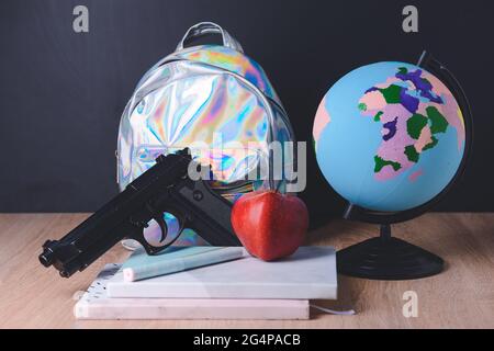 Pistole und Rucksack mit Schreibwaren auf dem Tisch im Klassenzimmer. Konzept des Schussdrehens Stockfoto