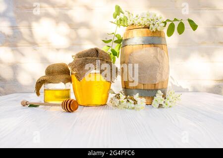 Floraler Bio-Honig in Holzfass und Glas mit frischen Akazienblüten auf dem Hintergrund der weißen Ziegelmauer mit einem Schatten von Bäumen. Stockfoto