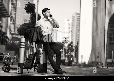 Junger asiatischer Geschäftsmann, der neben seinem Elektroroller auf der Straße steht und einen Anruf mit dem Handy im Schlepptau der modernen Stadt, schwarz-weiß, abgibt Stockfoto