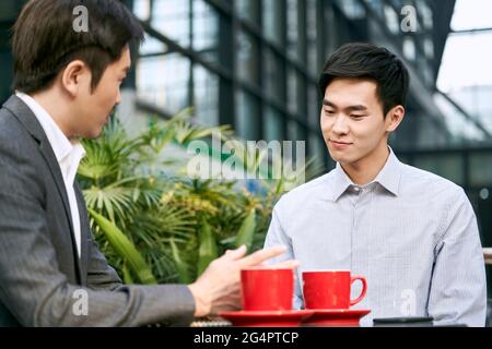 Zwei asiatische Geschäftsleute, die bei einem Kaffee im Freien über Geschäfte diskutieren Kaufen Stockfoto