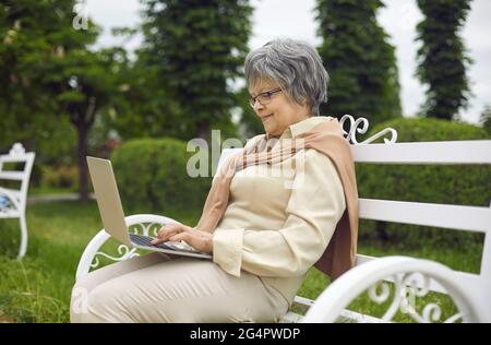 Alte Dame, die einen Laptop benutzt und sich im Park ausruht, während sie auf der Bank sitzt Stockfoto