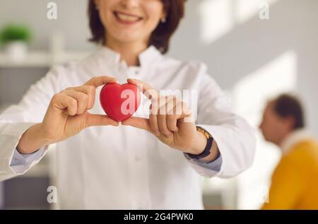 Nahaufnahme eines kleinen roten Herzens, das in den Händen einer lächelnden Kardiologin gehalten wird. Stockfoto