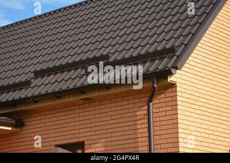 Eine Nahaufnahme auf einem metallbraunen Dach eines Backsteinhauses mit Schneestoppern, Schneeräumen, einer Regenrinne, einem Downauslauf, Soffit und Faszienbrett. Stockfoto