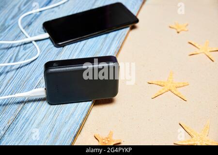 Powerbank lädt ein Smartphone auf einem Sommerhintergrund aus Holzbrettern und Sand mit Seesternen Stockfoto