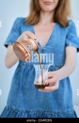 Nicht erkennbare Frau, die Kaffee aus dem Cazve in ein Glas gießt (Fokus auf Vordergrund) Stockfoto