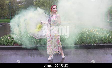 Entzückendes Mädchen in einem Kleid mit einem Blumendruck mit Regenbogen Zöpfen und Make-up. Tanzt mit einem Regenschirm, der sich in gelbem Rauch vor der Kulisse von FL Stockfoto