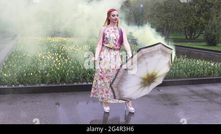 Entzückendes Mädchen in einem Kleid mit einem Blumendruck mit Regenbogen Zöpfen und Make-up. Tanzt mit einem Regenschirm, der sich in gelbem Rauch vor der Kulisse von FL Stockfoto