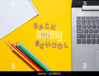 Zurück zur Schule minimalistisches Konzept. Laptop, Farbstifte, Notizbuch auf gelbem Hintergrund. Slogan Zurück in die Schule mit Holzbuchstaben Stockfoto