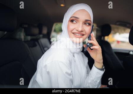 Schöne junge Frau im Hijab sitzt in einem Auto und spricht am Telefon Stockfoto