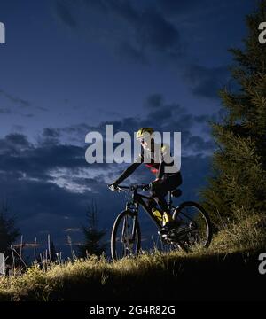 Lächelnder junger Mann, der mit dem Fahrrad bergab fährt, mit dem wunderschönen blauen Abendhimmel im Hintergrund. Fahrradfahrer im Sport Radfahren Anzug Radfahren hinunter grasbewachsenen Hügel in der Nacht. Konzept von Sport, Radfahren und aktiver Freizeit Stockfoto