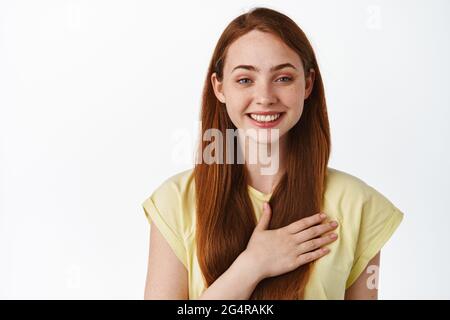 Nahaufnahme Porträt einer niedlichen lächelnden jungen Frau, halten Sie die Hand auf das Herz und schauen glücklich, sagen Sie danke, fühlen Sie sich erfreut und dankbar, gegen Weiß stehen Stockfoto