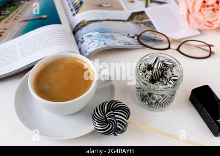 Tasse Kaffee mit wirbelschwarzem und weißem Lollipop, Süßigkeiten im Glasgefäß; eröffnete Zeitschrift mit Sommerseebildern und Gläsern auf einem Tisch Stockfoto