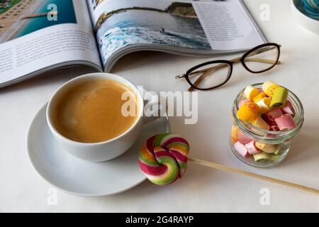 Tasse Kaffee mit buntem Lutscher, Bonbons in einem Glasglas; geöffnetes Magazin mit Sommerseebildern und Gläsern auf einem Tisch Stockfoto