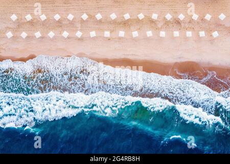 Luftaufnahme des schönen Strandes mit weißen Sonnenschirmen und blauem Meer mit großen Wellen. Stockfoto