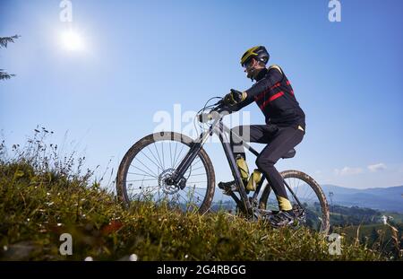 Junger Mann mit Schutzhelm und Brille, der mit blauem Himmel auf dem Hintergrund bergauf radelt. Männlicher Radfahrer im Radanzug bergauf auf auf dem Mountainbike klettern. Konzept von Sport, Mountainbiken und aktiver Freizeit Stockfoto