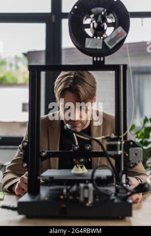 Fokussierter junger Designer, der sich mit dem 3D-Drucker beschäftigt, der im modernen Büro ein Modell herstellt Stockfoto