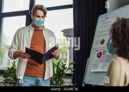 Junger Mann in medizinischer Maske, der Ordner mit Papieren in der Nähe des Flipchart hält, und afroamerikanischer Kollege im Büro Stockfoto