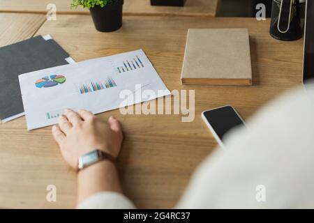 Teilansicht der männlichen Hand in der Armbanduhr, die Papier berührt, mit Grafiken und Diagrammen in der Nähe des Mobiltelefons auf dem Holzschreibtisch Stockfoto