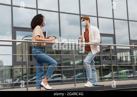 Junges interrassisches Paar in medizinischen Masken und legerer Kleidung, das mit Pappbechern in den Händen in der Nähe des Glasgebäudes auf der Straße redet Stockfoto