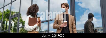 Junge interrassische Kollegen in medizinischen Masken gehen mit Pappbechern und Dokumenten in der Nähe von Glasgebäude, Banner Stockfoto