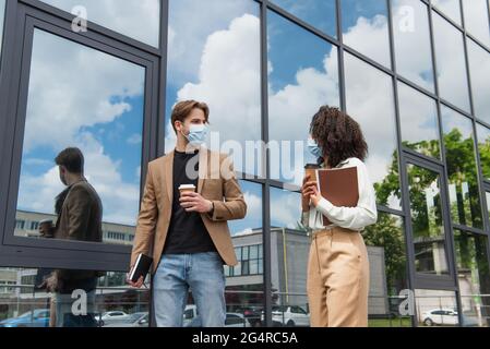 Junge interrassische Kollegen in medizinischen Masken, die mit Kaffeetassen und Dokumenten in der Nähe des modernen Gebäudes spazieren Stockfoto