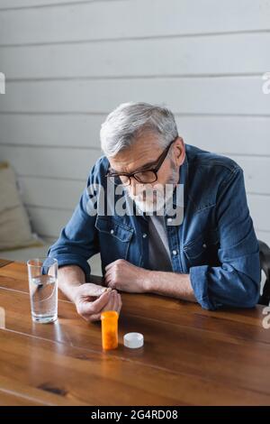 Älterer Mann in einer Brille, der die Pille in der Nähe eines Glases Wasser zu Hause ansieht Stockfoto