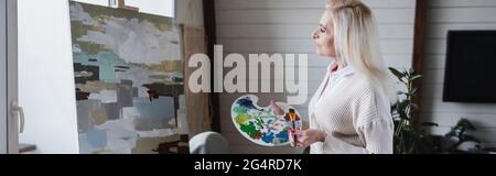 Seitenansicht einer älteren Frau mit Palette und Pinsel in der Nähe von Malerei, Banner Stockfoto