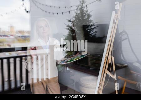 Eine ältere Frau hält zu Hause die Palette in der Nähe der Staffelei und des Fensters Stockfoto