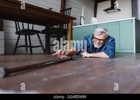 Oberflächenhöhe des behinderten älteren Mannes, der zu Hause auf dem Boden liegt und Krücke nimmt Stockfoto
