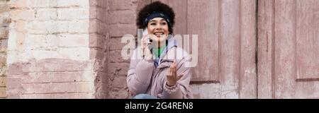 Fröhliche afroamerikanische Frau, die in der Nähe des Gebäudes auf dem Handy spricht, Spruchband Stockfoto