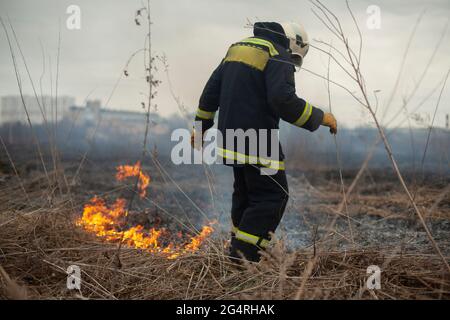 Ein Feuerwehrmann löscht trockenes Gras. Ein Feuerwehrmann bekämpft ein Feuer in einem offenen Bereich. Rettungsaktionen gegen Flammen. Ein ökologischer Katastrophenbrand Stockfoto