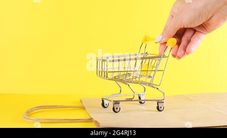 Weibliche Hand schiebt ein Spielzeug Warenkorb auf gelbem Hintergrund, Nahaufnahme. Black Friday Shopping- und Rabattkonzept Stockfoto