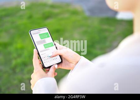 Frau schreibt mit Telefon im Freien. SMS mit dem Smartphone. Digitale sms und Instant Messaging-Chat. Person, die im Park vor dem Haus ein Mobiltelefon benutzt. Stockfoto