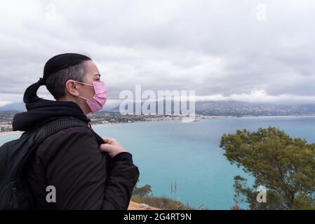 Frau mit kurzen Haaren in schwarz gekleidet und trägt einen Rucksack und trägt eine rosa Schutzmaske für Covid, blickt über die mediterrane Meereslandschaft Stockfoto