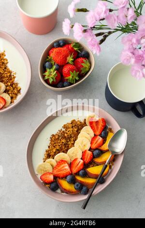 Leckeres gesundes Frühstück, Müsli mit Joghurt und süßen Früchten, Banane und Erdbeeren, Blaubeeren und Pfirsiche, Milch im Becher und Blumen Stockfoto