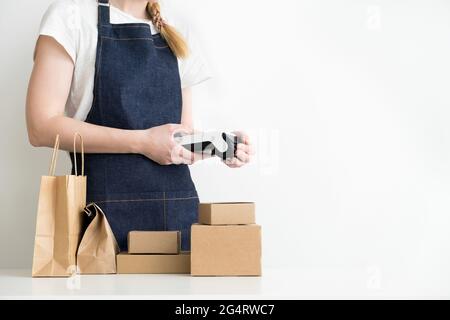 Frau, die ein modernes Zahlungs-POS-Terminal in Händen hält, mit Kartons, Pappverpackungen und Kraftbeutel auf dem Tisch. Konzept für kleine Unternehmen. Stockfoto