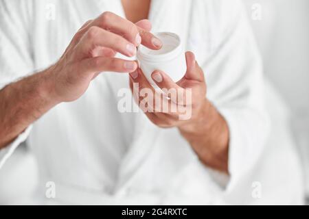 Mann im Bademantel nimmt Creme aus offenem Glas auf hellem Hintergrund Nahaufnahme Stockfoto