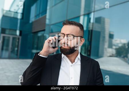 Porträt eines ernsthaften Geschäftsmannes im Anzug, der auf dem Mobiltelefon spricht, während er in der Nähe des Bürozentrums läuft und Platz zum Kopieren verwendet. Schöner bärtiger Mann, der sich untersprach Stockfoto