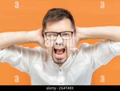 Porträt eines gefühlsmäßig nervösen bärtigen Mannes in klassischem weißen Hemd vor einem orangefarbenen Hintergrund, isoliert, die Ohren mit Händen und Schreien bedeckt. Kopie sp Stockfoto