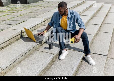 Junge konzentriert stilvolle afroamerikanische freiberufliche Geschäftsmann männlich arbeiten mit Laptop und Handy im Freien, auf der Treppe sitzen Stockfoto