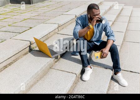 Junge attraktive afroamerikanische freiberufliche Geschäftsmann oder Student sitzen auf Treppen im städtischen Bereich und tun einige Arbeit auf Laptop. Gutaussehender Mann nehmen Stockfoto