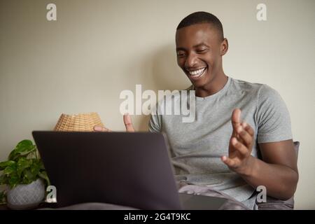 Lachender junger afrikanischer Mann, der zu Hause einen Laptop im Bett benutzt Stockfoto