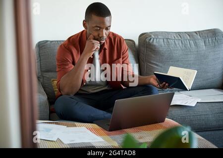 Junger afrikanischer Mann, der von zu Hause aus auf seinem Sofa arbeitet Stockfoto