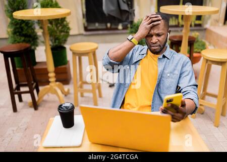 Junge müde Afro-amerikanische Freiberufler oder Student sitzen am Kaffeetisch mit Laptop und Smartphone und berühren seinen Kopf denken, Probleme mit Stockfoto