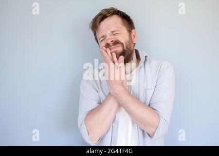 Der junge Europäer im Hemd leidet unter schweren Zahnschmerzen, er hält die Hand an der Wange, schließt die Augen und grimaces. Medizinisches Konzept. Stockfoto